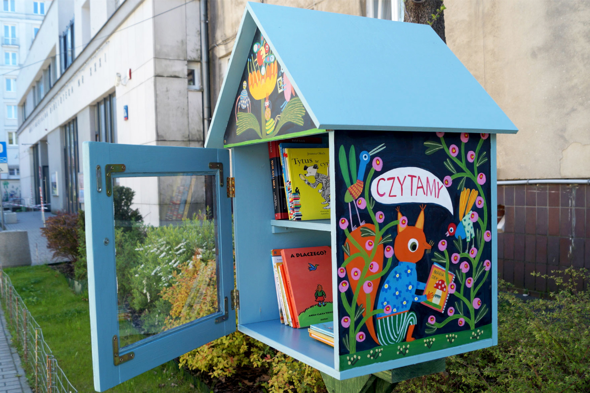 Niebieska plenerowa biblioteczka w kształcie małego domku, stojąca przy ulicy. Drzwiczki są otwarte, a w środku widać kolorowe książki dla dzieci. Na bocznej ścianie domku znajduje się barwna ilustracja z leśnymi zwierzętami i napisem ‘Czytamy’. W tle widać budynek biblioteki oraz roślinność.